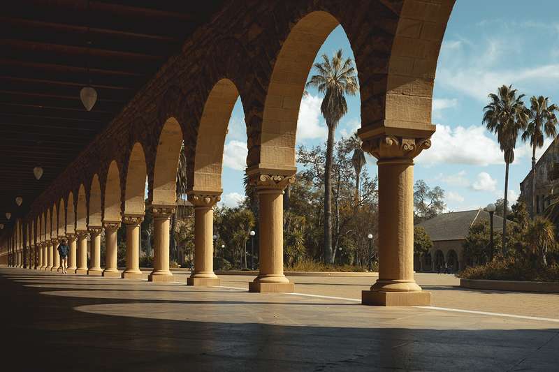 A view through an arched sandstone arcade looking out toward the Main Quad on the Stanford campus, with palm trees and varied plantings against a partly cloudy blue sky.