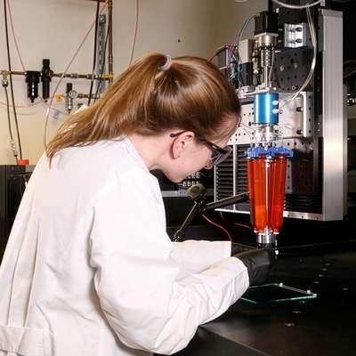 A researcher in a lab coat operates a 3D printing device that deposits red material through a nozzle onto a glass plate.