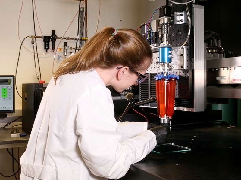 A researcher in a lab coat operates a 3D printing device that deposits red material through a nozzle onto a glass plate.