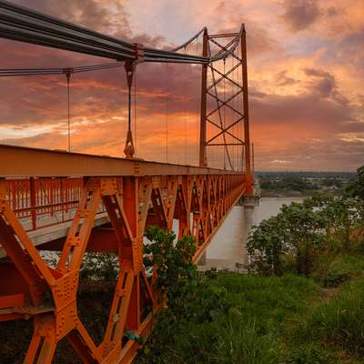 An orange steel suspension bridge stretches over a wide river at sunset beneath a vivid orange and pink sky.