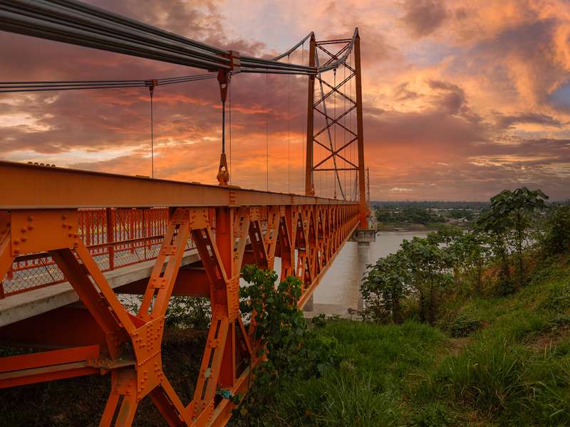 An orange steel suspension bridge stretches over a wide river at sunset beneath a vivid orange and pink sky.