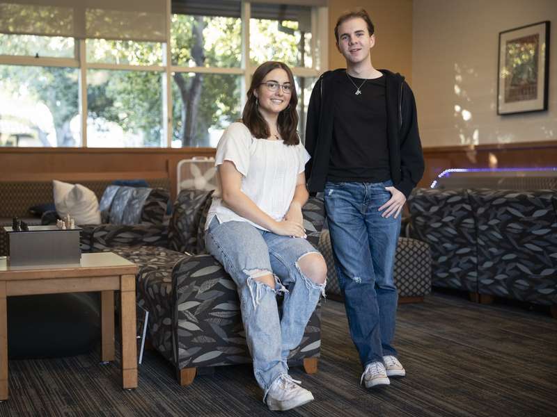 Stella Vangelis and Peter Bennett pose together in a casual campus lounge, one seated and one standing, with sunlit trees visible through the windows behind them.