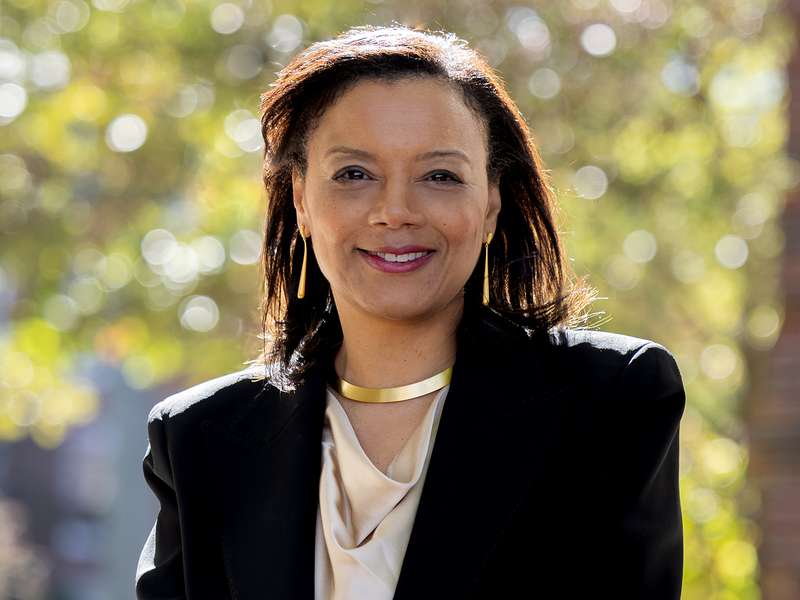 Woman in a black blazer and gold necklace smiling outdoors with blurred trees in the background.
