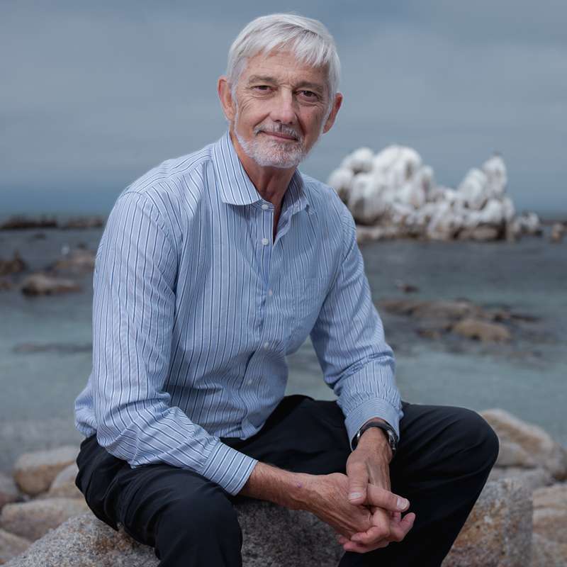 Stephen Palumbi sitting on rocks near the ocean and posing for a photo.