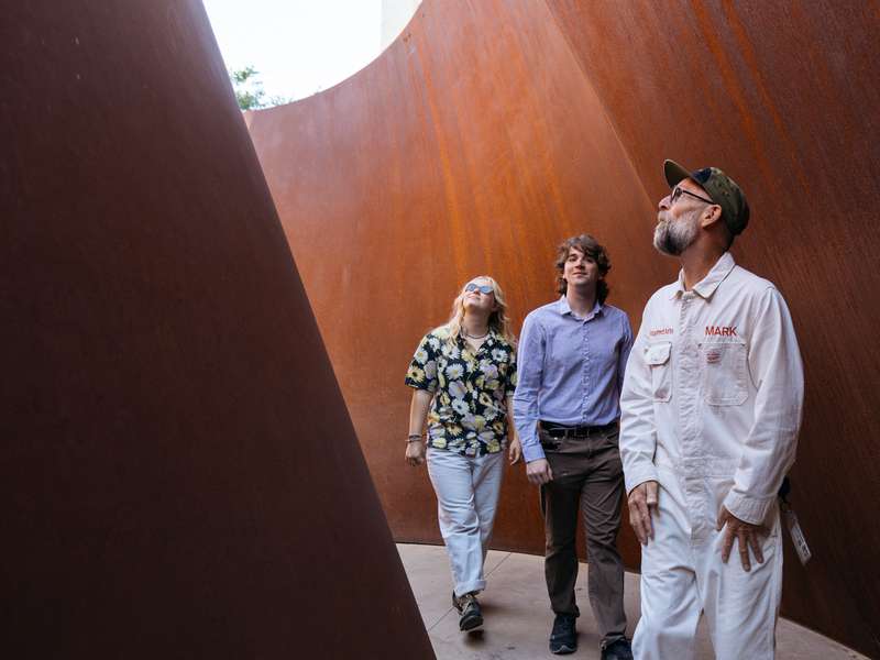 Two people and guide Mark Shunney in white coveralls walk between the leaning rust-colored steel walls of a curving sculpture.