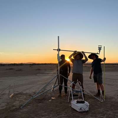 Researchers Adam Brandt, Josh Romo, and Taylor Adams set up a tripod-mounted weather sensor on a desert flat at sunset.