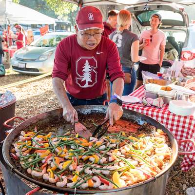 A person cooks a large dish with shrimp and vegetables at a lively tailgate party, with friends enjoying food in the background.