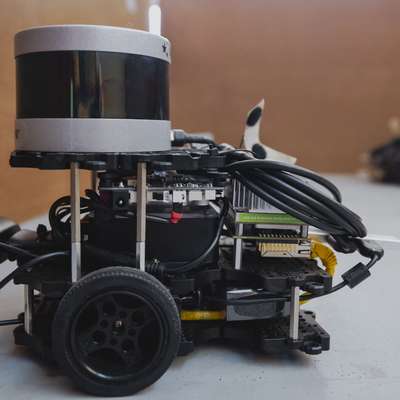 Small wheeled robot with a cylindrical lidar sensor, circuit boards and cables on a table, another robot blurred behind.