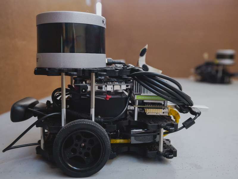 Small wheeled robot with a cylindrical lidar sensor, circuit boards and cables on a table, another robot blurred behind.