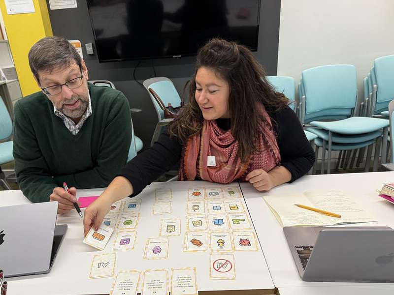 Two people collaborate at a table, examining cards and notes while using laptops in a classroom setting.