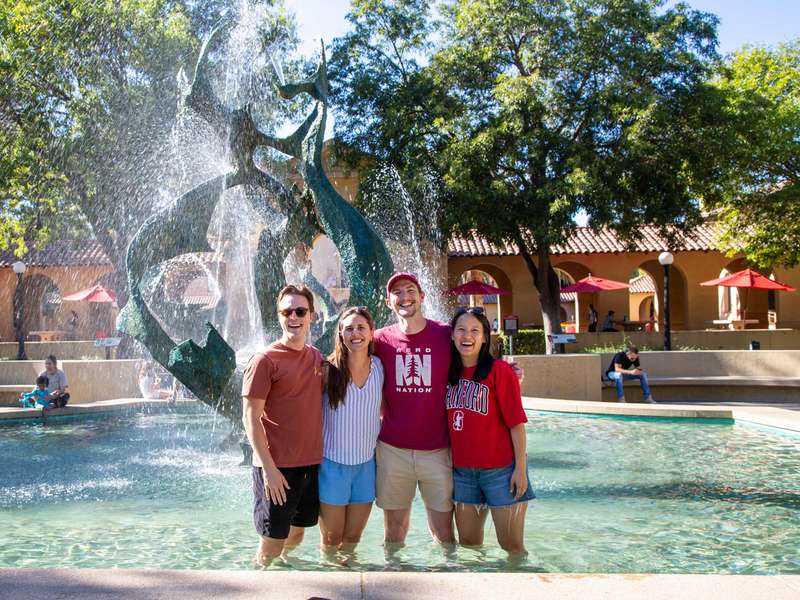 Four friends stand arm in arm in a pool of water, smiling as water sprays from White Memorial Fountain behind them.