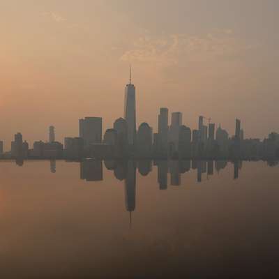 The Manhattan skyline sits under a haze at sunrise, its silhouette mirrored in the still water below.