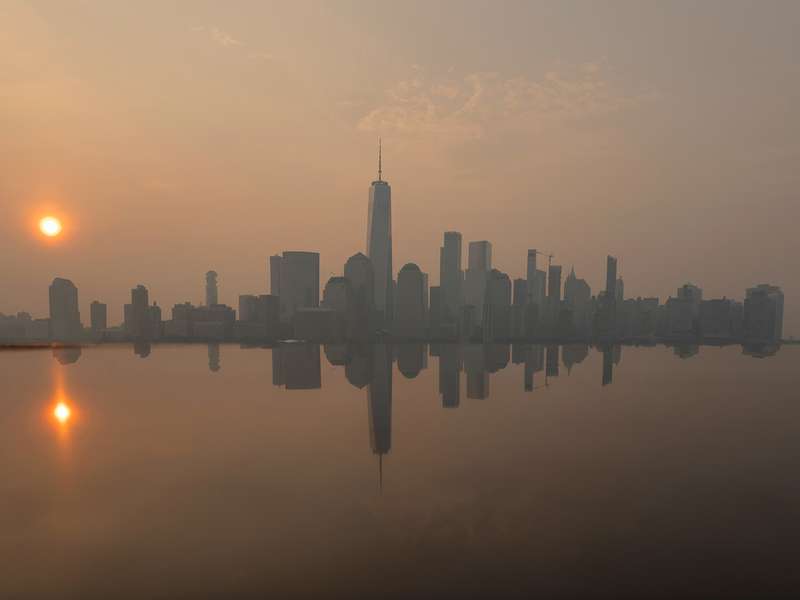 The Manhattan skyline sits under a haze at sunrise, its silhouette mirrored in the still water below.