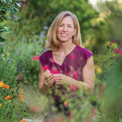 Sattely with long blond hair in purple top smiles while standing in a colorful flower garden.