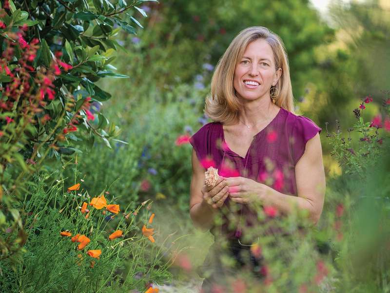 Sattely with long blond hair in purple top smiles while standing in a colorful flower garden.