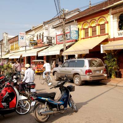 A busy street scene in a market with shops and restaurants, featuring parked motorcycles and people walking and cycling along the road.