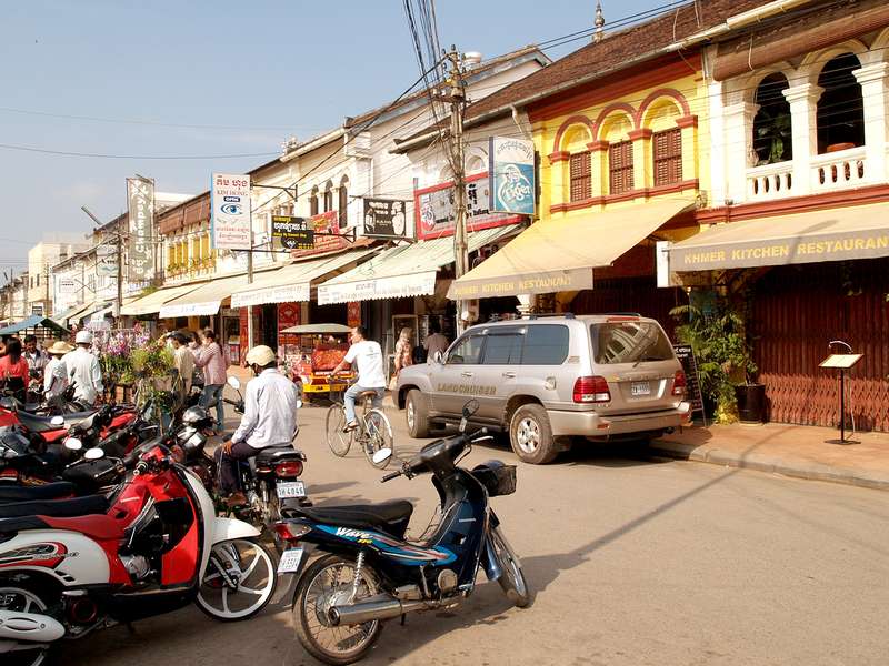 A busy street scene in a market with shops and restaurants, featuring parked motorcycles and people walking and cycling along the road.
