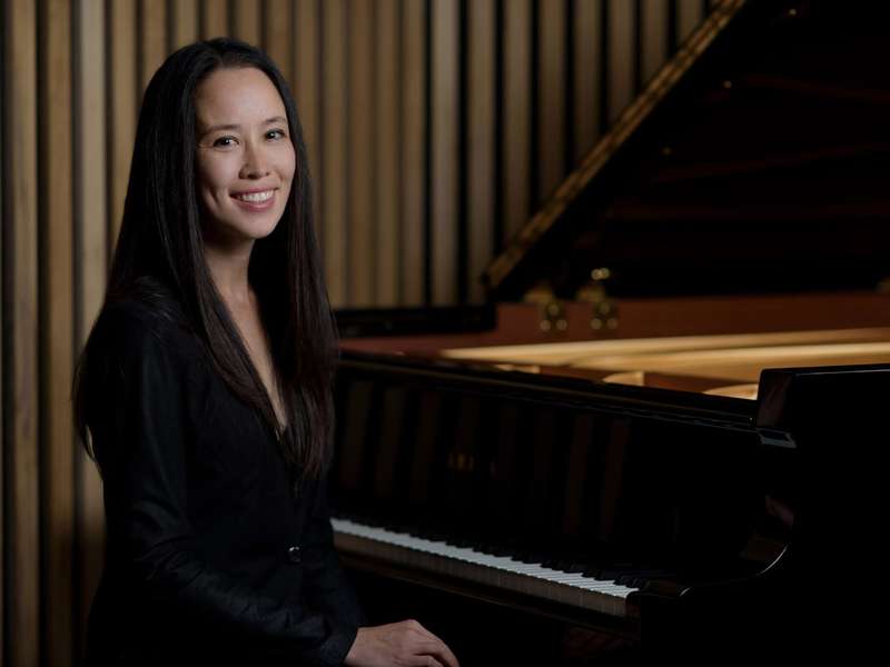 Pianist smiles warmly while sitting beside a grand Yamaha piano in a wooden-paneled room.