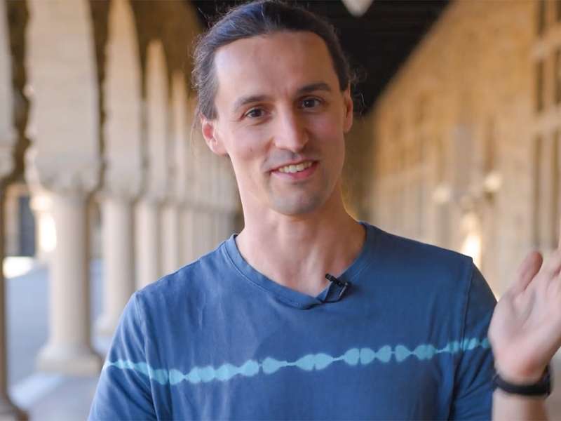 Ettore Biondi, wearing a clip-on microphone, waves gestures with his hands while talking in a sunlit sandstone arcade.