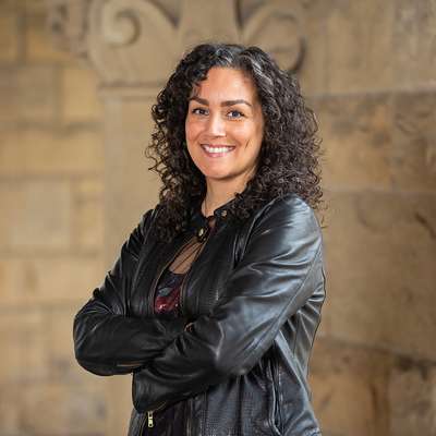 Rose Salseda posing and smiling for a profile photo and wearing a black leather jacket while standing with folded arms before a stone wall.