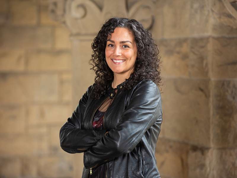 Rose Salseda posing and smiling for a profile photo and wearing a black leather jacket while standing with folded arms before a stone wall.
