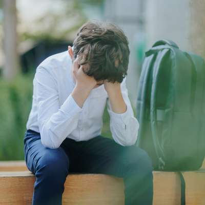 A young boy sits on a bench with his head in his hands, looking distressed, with a backpack beside him.