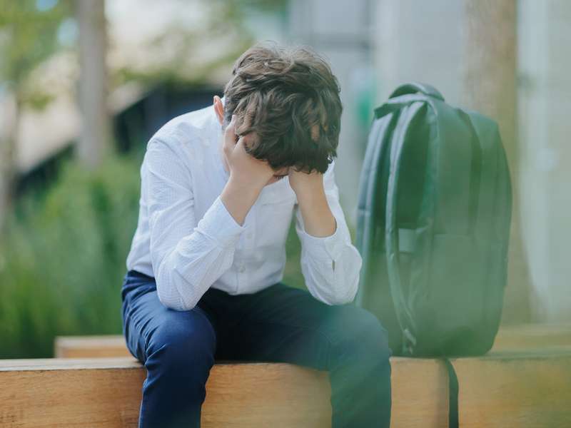 A young boy sits on a bench with his head in his hands, looking distressed, with a backpack beside him.