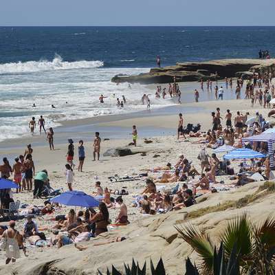 Crowds sunbathe and swim at a busy beach lined with rocks and colorful umbrellas.