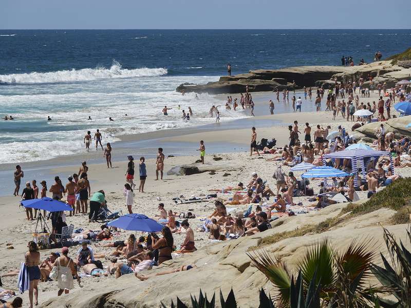 Crowds sunbathe and swim at a busy beach lined with rocks and colorful umbrellas.