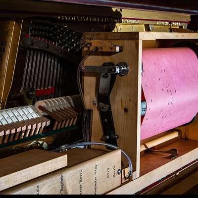 A player piano's mechanism holds a pink piano roll on its spool, with hammers and strings visible inside the open piano case.