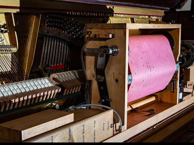 A player piano's mechanism holds a pink piano roll on its spool, with hammers and strings visible inside the open piano case.