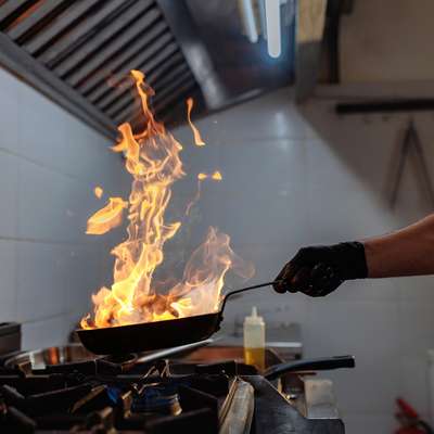 A gloved chef holds a flaming pan over a gas stove's blue flame in a restaurant kitchen.