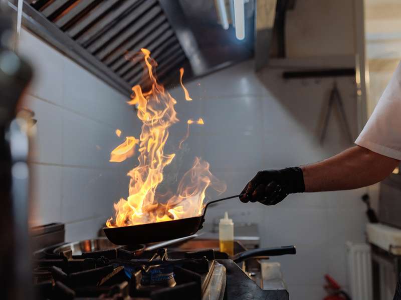 A gloved chef holds a flaming pan over a gas stove's blue flame in a restaurant kitchen.