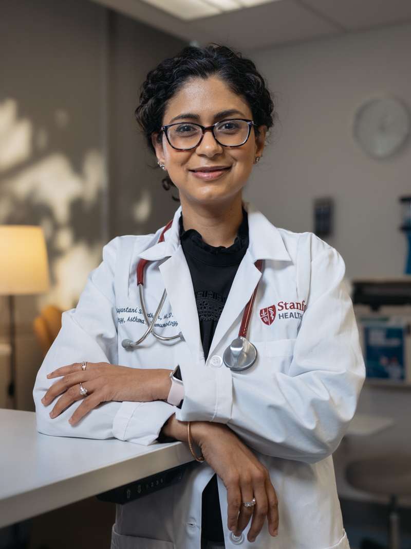 Tina Sindher in a lab coat stands in a clinic, showcasing a patient care environment.