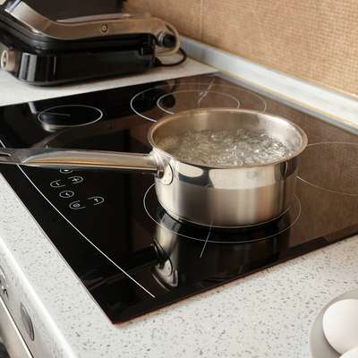 A metal pot filled with boiling water sits on a modern stovetop near eggs and tomatoes on a plate.
