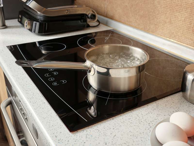 A metal pot filled with boiling water sits on a modern stovetop near eggs and tomatoes on a plate.