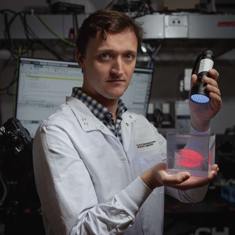 Mark Skylar-Scott faces the camera in a lab setting, holding a clear container with a glowing red biological specimen in one hand and a UV flashlight raised in the other.