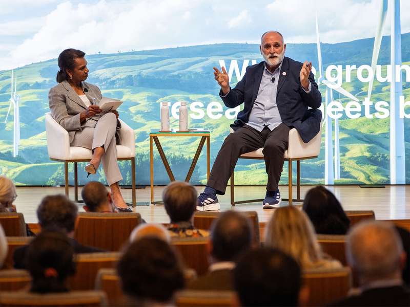 A panel discussion featuring Condoleezza Rice and Jose Andres on stage with a green landscape backdrop and wind turbines, engaging an audience.