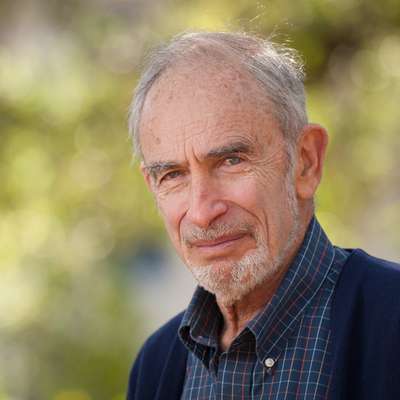 Paul Ehrlich posing for a photo outdoors, wearing a plaid shirt and dark sweater, surrounded by a soft, blurred green background.