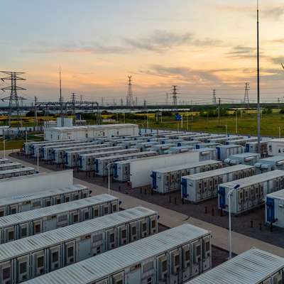 Rows of white container-sized units at an energy storage facility, with power lines and a wind turbine in the distance at sunset.
