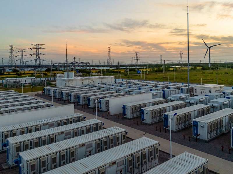 Rows of white container-sized units at an energy storage facility, with power lines and a wind turbine in the distance at sunset.