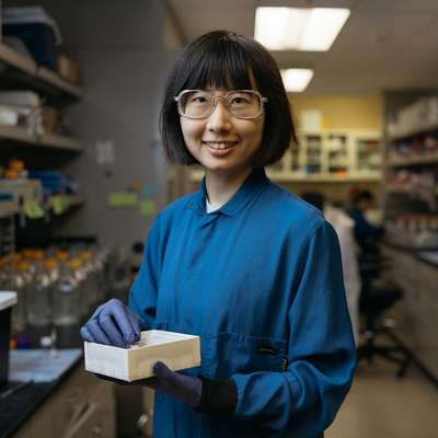 Nina Zhao in safety glasses and a blue lab coat smiles while holding a box of samples in a lab aisle lined with shelves of supplies.