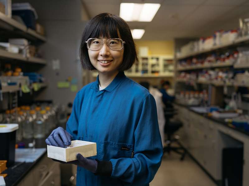 Nina Zhao in safety glasses and a blue lab coat smiles while holding a box of samples in a lab aisle lined with shelves of supplies.