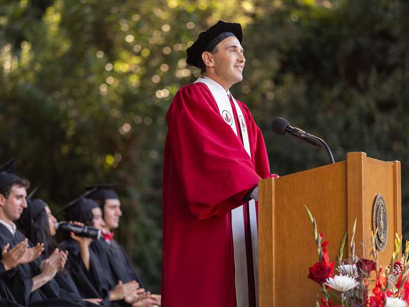 President Levin in crimson academic robe and black tam speaks at a podium while seated students in black gowns applaud.