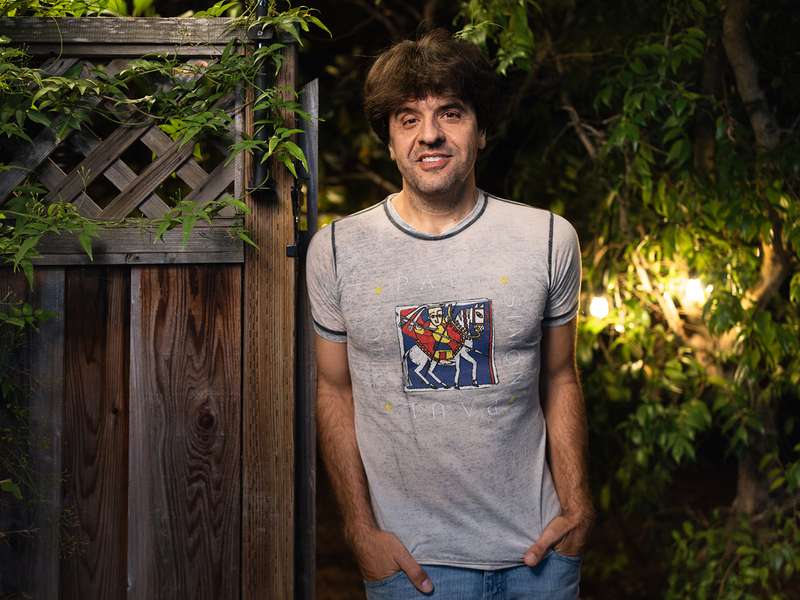 Karl Deisseroth stands with his hands in his pockets beside a wooden garden gate at night, with string lights glowing behind him.