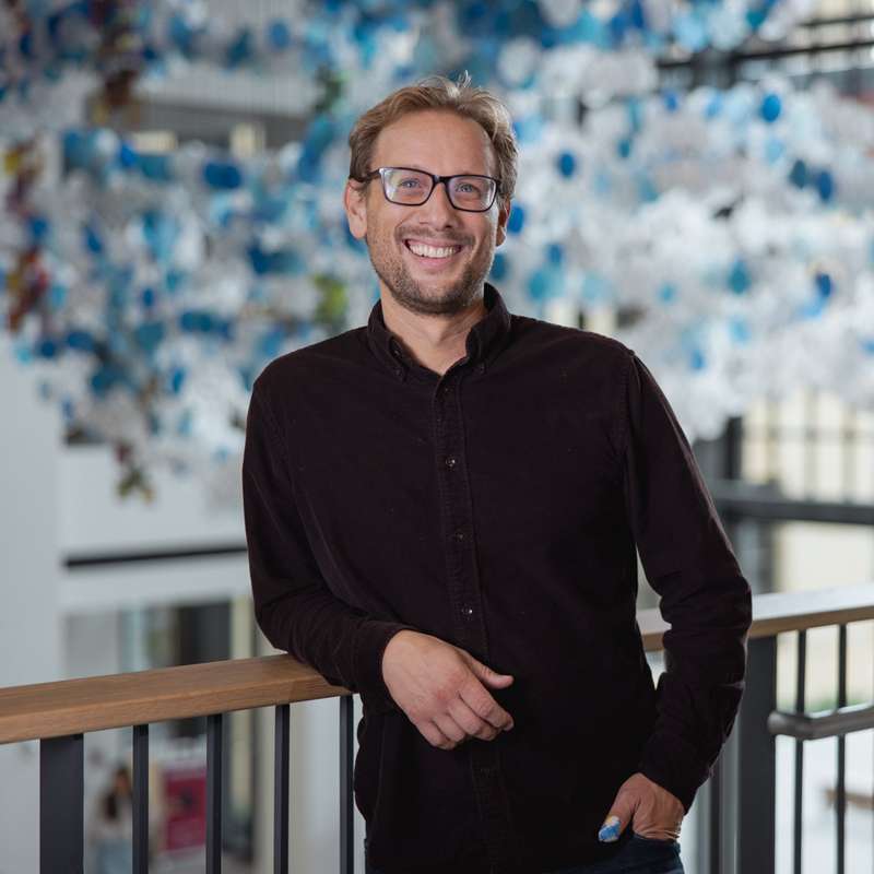 Jason Yeatman smiling and leaning on a railing indoors, against a blurred blue and white hanging art installation in the background.