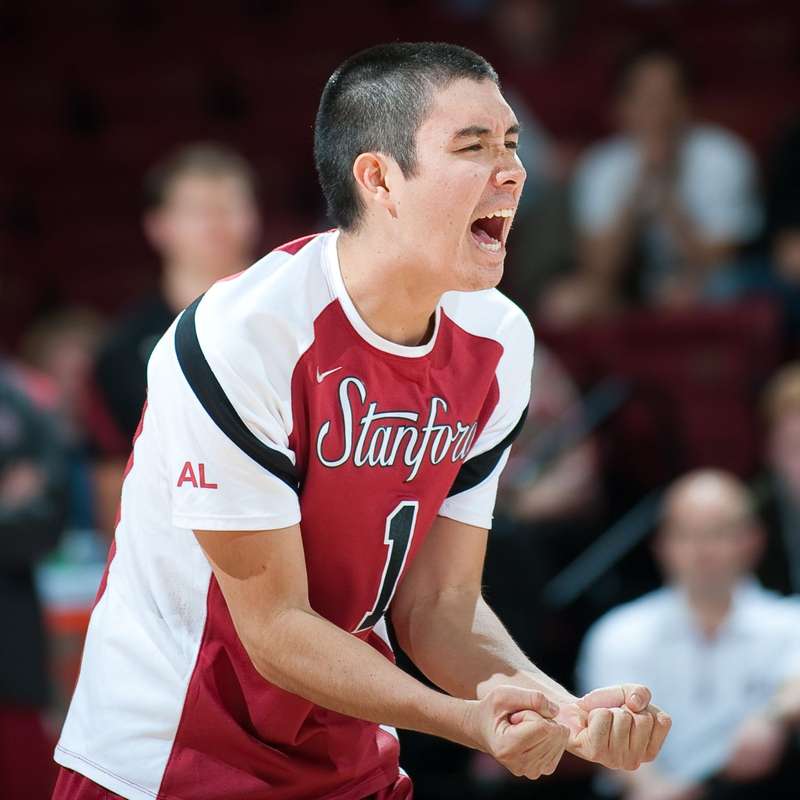 Erik Shoji celebrates enthusiastically during a volleyball match with spectators blurred out in the background.