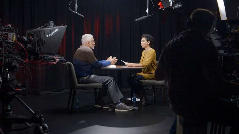 Stanford historian Clayborne Carson and Zainab Taymuree engage in a conversation at a round table during a filmed interview, surrounded by cameras and studio equipment.