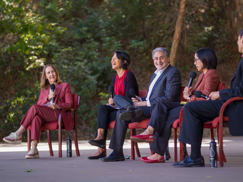 Stanford faculty members sit for a panel discussion, laughing together while holding microphones outdoors.