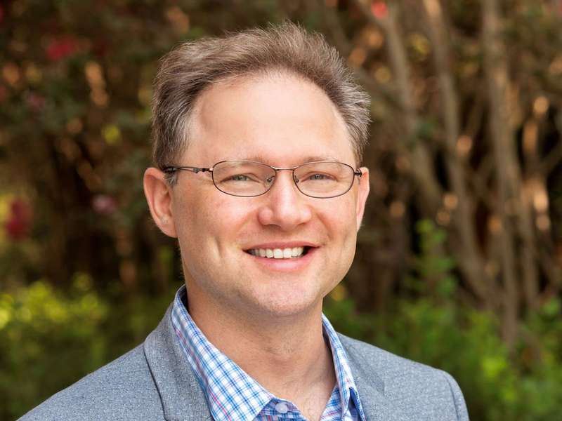A smiling man with glasses wears a gray blazer over a checked shirt, with green foliage and flowers in the background.
